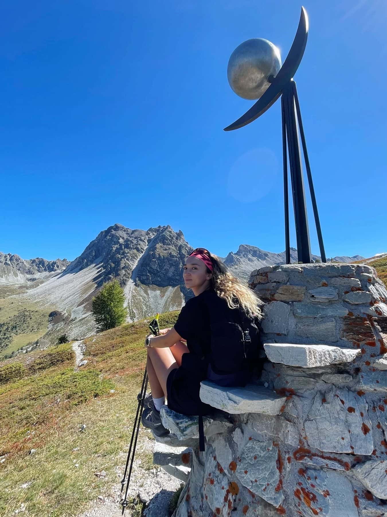 Lisa assise sur un muret de pierre au pied d'une sculpture, au-dessus du val d'Anniviers